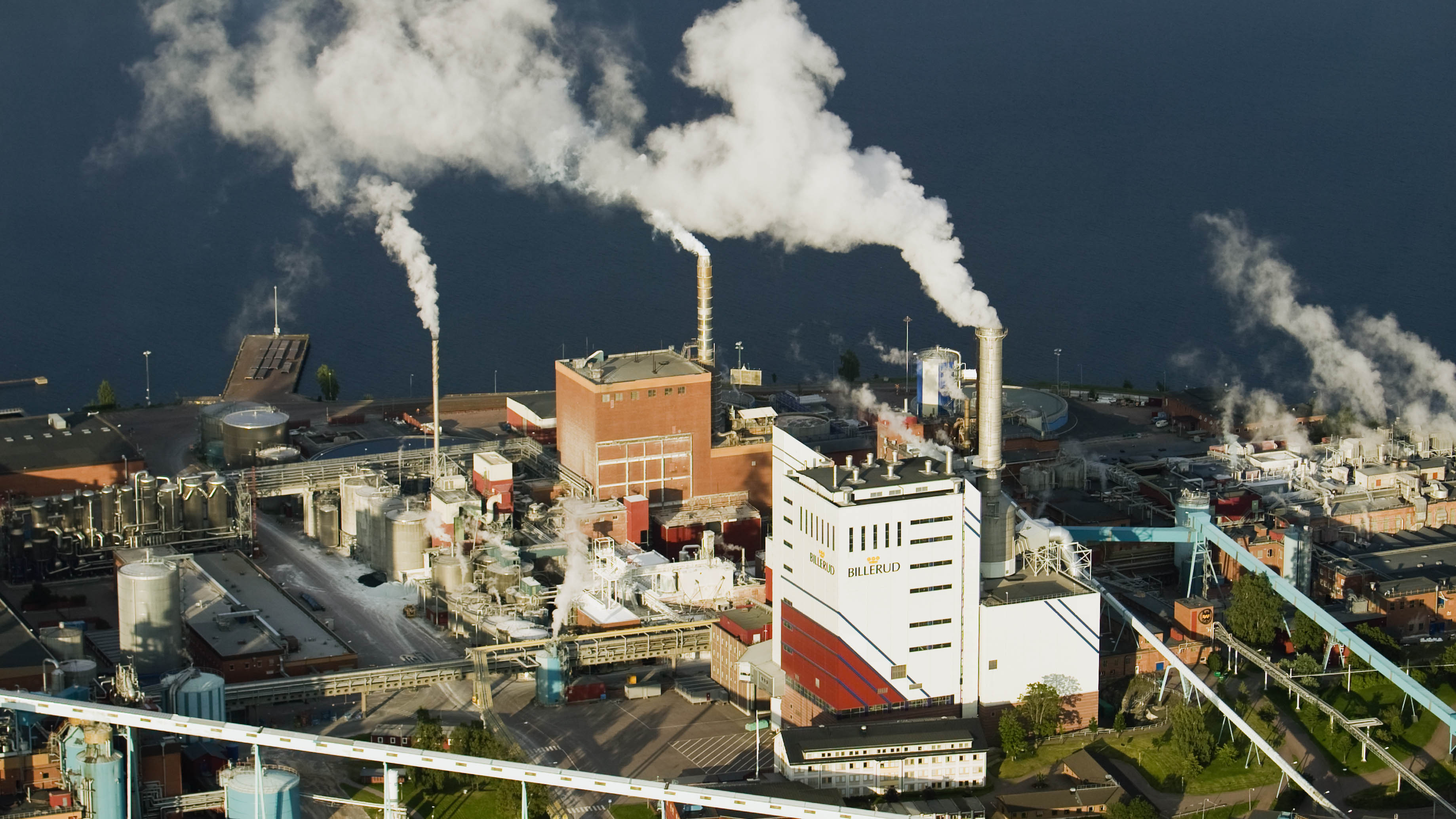 Fotograf: Roine Magnusson
Bildbyrå: Johnér Flygfoto över fabrik med vit rök bolmande ur skorstenarna. I bakgrunden en sjö..