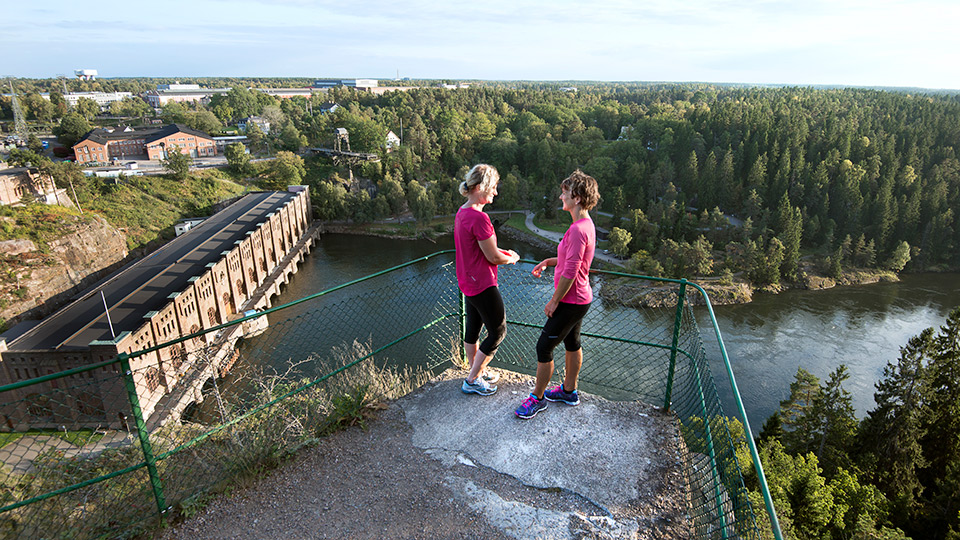 Två kvinnor samtalar vid en utkikspunkt med vatten nedanför och skog och byggnader i bakgrunden
