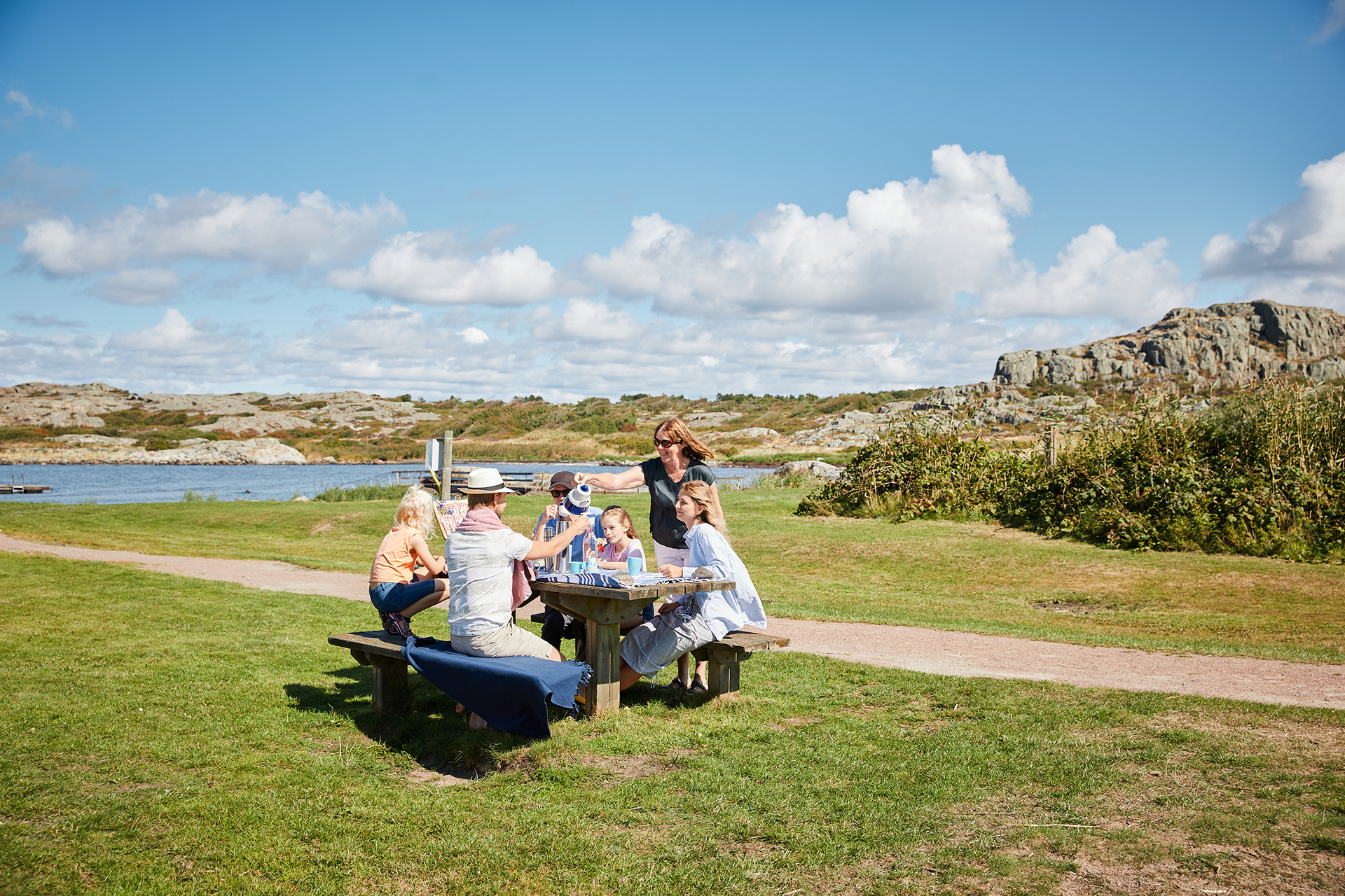Fotograf: Öckerö kommun Två vuxna och fyra barn som sitter runt ett picknickbord i skärgårdsmiljö