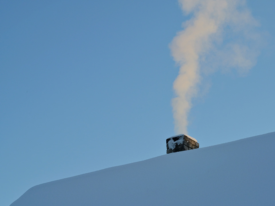 Fotograf: Andres Siimon
Bildbyrå: Unsplash Snötäckt tak med en rykande skorsten mot en blå himmel