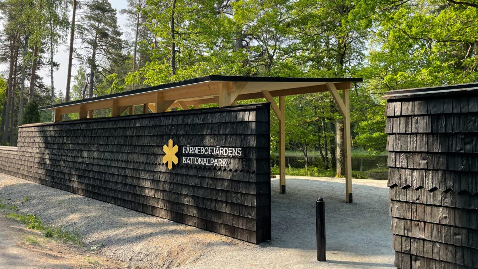 Fotograf: Länsstyrelsen Gävleborg
Bildbyrå: Länsstyrelsen Gävleborg Huvudentré Nord i Färnebofjärdens nationalpark med en skylt på en mörk trävägg, omgiven av grönskande träd.